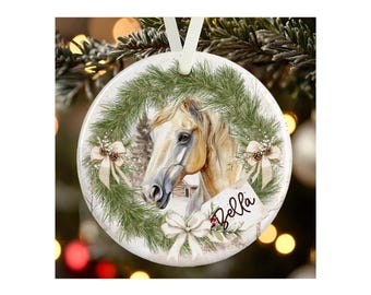 May include: A round white ceramic ornament featuring a printed illustration of a light-colored horse centered within a green pine wreath. The design includes three cream-colored bows and a small tag with the name Bella written in black script.