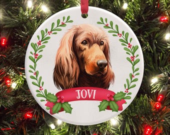 May include: A round white ceramic ornament featuring a portrait of a brown Irish Setter dog. The dog is framed by a green holly wreath with red berries. A red ribbon banner across the bottom displays the name JOVI in white capital letters.