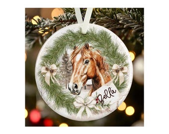May include: A round white ceramic ornament featuring a brown horse head centered within a green pine wreath. The design includes three cream-colored bows and a small rectangular tag with the name Bella written in black cursive script.