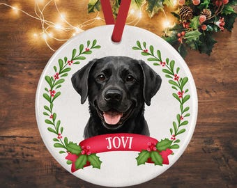 May include: A round white ceramic ornament featuring a black Labrador retriever portrait centered within a green holly wreath with red berries. A red ribbon banner across the bottom displays the name JOVI in white capital letters. A red ribbon loop is attached.