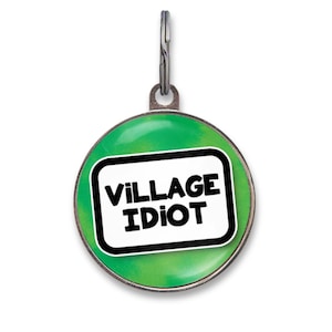May include: A round metal pet ID tag with a green background and a white rectangular center featuring the black text VILLAGE IDIOT. The tag includes a silver-toned metal border and a small metal ring at the top for attachment to a collar.