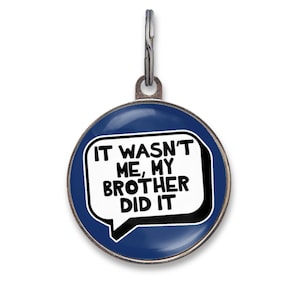 May include: A round, dark blue pet ID tag with a silver-toned metal rim and a metal split ring at the top. The center features a white speech bubble graphic with black text that reads, IT WASN'T ME, MY BROTHER DID IT, in a bold, sans-serif font.