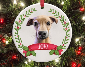 May include: A round white ceramic ornament featuring a tan dog illustration centered within a green holly and red berry wreath. A red ribbon banner across the bottom displays the name JOVI in white capital letters. A red ribbon loop hangs from the top.