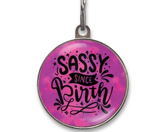 May include: A round metal pet ID tag featuring a vibrant pink and purple tie-dye background. The center displays black stylized lettering that reads SASSY SINCE BIRTH, accented with small decorative stars and flourishes, attached to a metal ring.