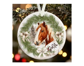 May include: A round white ceramic ornament featuring a brown and white horse head centered within a green pine wreath. The wreath is decorated with three white bows and small pinecones. The name Bella is printed in black script on a white tag.