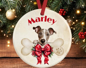 May include: A round, cream-colored ceramic ornament featuring a brown and white dog peeking over a large bone. The name Marley is printed in red above the dog. A large red bow with green holly and red berries sits below the bone, and a paw print is visible on the right side of the bone.