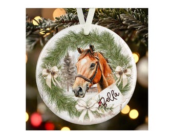 May include: A round white ceramic ornament featuring a brown horse head centered within a green pine wreath. The wreath is decorated with three cream-colored bows and pinecones. A white tag with the name Bella in black script is attached.