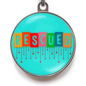 May include: A round, turquoise blue pet tag with a silver border. The word "RESCUED" is spelled out in colorful blocks with red hearts hanging from each letter.