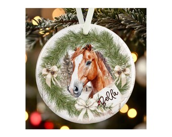 May include: A round white ceramic ornament featuring a brown horse with a white blaze on its face, framed by a green pine wreath with white bows and pinecones. The name Bella is printed in black script on a white tag attached to the wreath.
