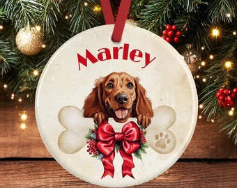 Op de afbeelding: Een ronde keramische kerstornament met een bruine hond die zijn poten op een groot wit bot laat rusten. De naam Marley is in het rood boven de hond gedrukt. Een rode strik met groene hulst en rode bessen is in het midden onder de poten geplaatst.