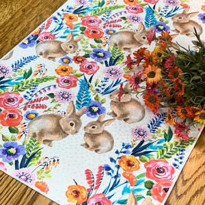 May include: A white table runner with a floral pattern featuring bunnies, flowers, and leaves in shades of pink, orange, yellow, blue, and green. The background is a light cream colour with a subtle polka dot pattern.