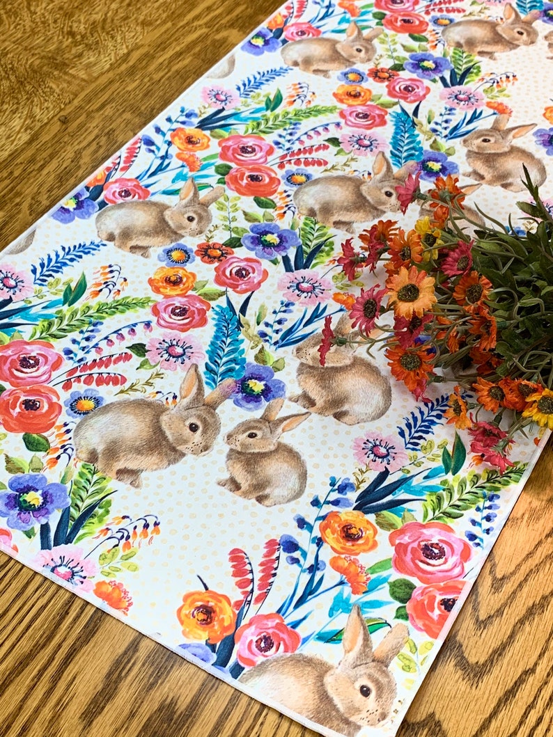 May include: A white table runner with a floral pattern featuring bunnies, flowers, and leaves in shades of pink, orange, yellow, blue, and green. The background is a light cream colour with a subtle polka dot pattern.
