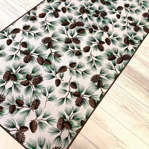 May include: A white runner rug with a brown and green pine cone pattern. The rug has a brown border.