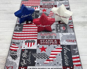 Puede incluir: Un camino de mesa patriótico con un diseño de la bandera estadounidense en gris, rojo y blanco. Tres decoraciones de estrellas de madera en azul, rojo y blanco se colocan encima. Las estrellas están inscritas con las palabras "Liberty", "America" y "Freedom".