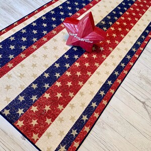 May include: A red, white, and blue American flag runner with glitter stars. The runner is on a light wood floor with a red wooden star that says "America" on it.