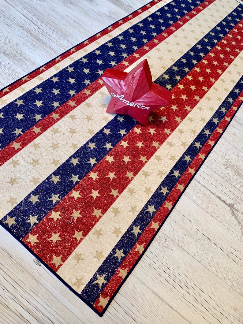 May include: A red, white, and blue American flag runner with glitter stars. The runner is on a light wood floor with a red wooden star that says "America" on it.