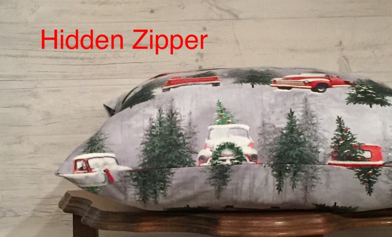 May include: A gray pillow with a Christmas-themed print featuring red trucks with Christmas trees on top, surrounded by green trees. The pillow has a hidden zipper.