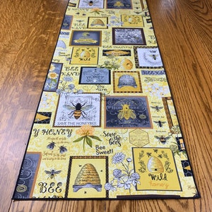 May include: A yellow and black table runner with a bee theme. The runner features a variety of bee-related images, including bees, honeycombs, and flowers. The text on the runner includes phrases like "Bee Kind", "Save the Honeybee", and "Wild Honey".