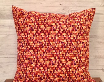 May include: A square throw pillow featuring a dense, repeating woven pattern in shades of deep purple, bright orange, and golden yellow. The fabric displays a textured, grid-like appearance with small, alternating rectangular shapes throughout the surface.
