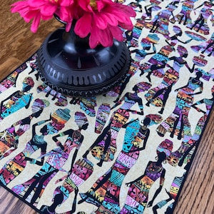 May include: A black and beige table runner with a repeating pattern of colorful silhouettes of women in traditional African clothing. The women are carrying baskets, jugs, and other items.
