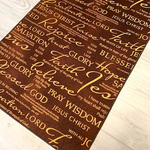May include: A rectangular brown rug with gold script featuring religious words like "Faith", "Glory", and "Jesus". The rug has a textured appearance and is placed on a light wood floor.