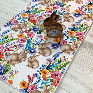 May include: A white table runner with a floral pattern featuring bunnies and the text "Easter Greetings". The floral pattern includes pink, red, orange, yellow, blue, and green flowers.