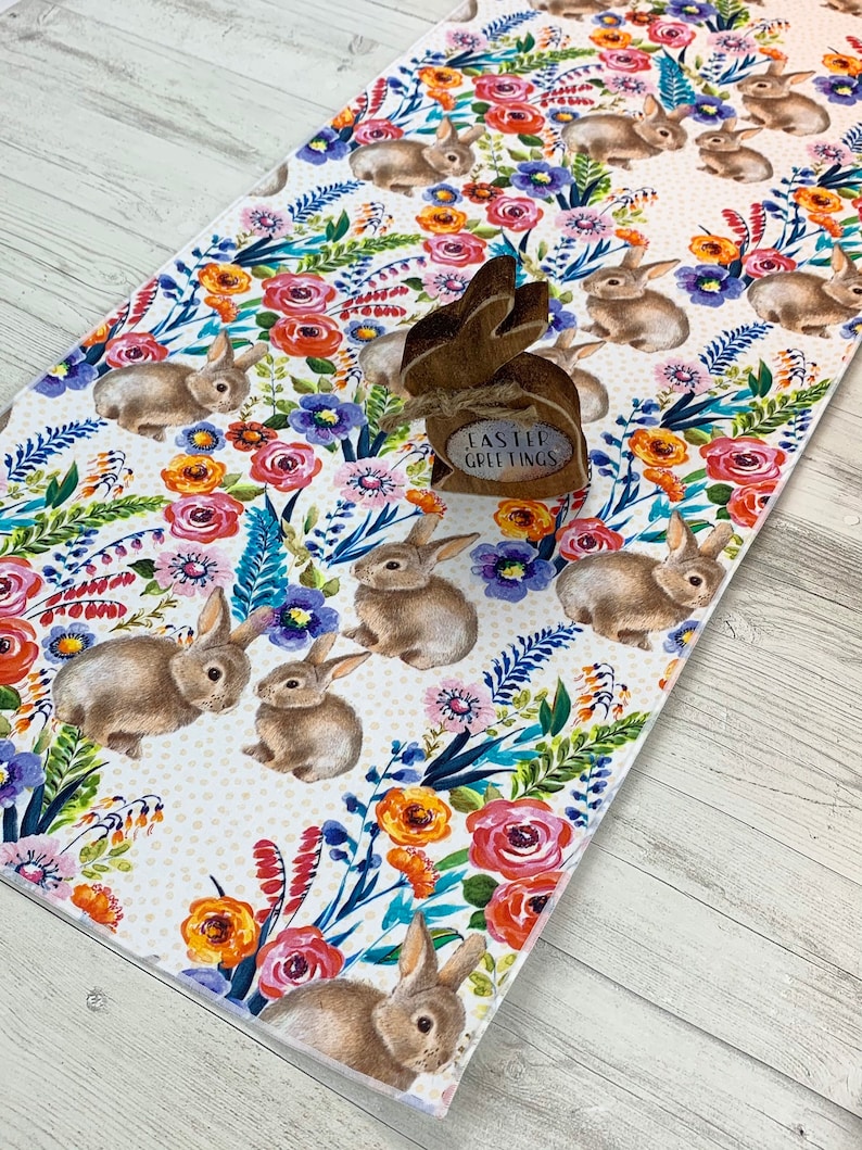 May include: A white table runner with a floral pattern featuring bunnies and the text "Easter Greetings". The floral pattern includes pink, red, orange, yellow, blue, and green flowers.