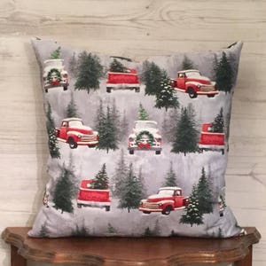 May include: A gray throw pillow with a festive Christmas design featuring red pickup trucks carrying Christmas trees against a snowy background.