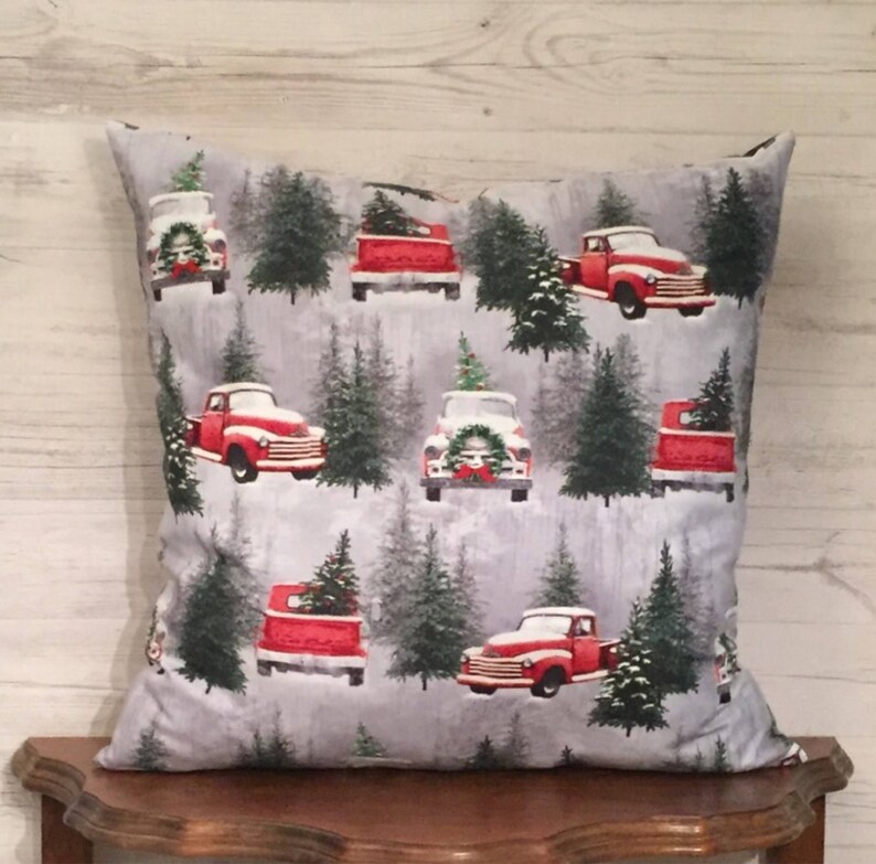 May include: A gray throw pillow with a festive Christmas design featuring red pickup trucks carrying Christmas trees against a snowy background.