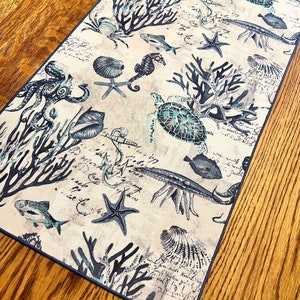 May include: A beige table runner with a blue and white nautical design. The runner features seahorses, starfish, seashells, coral, and fish. The background is a light beige with a faded script pattern.