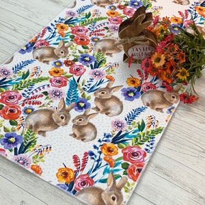 May include: A white table runner with a floral and bunny pattern. The runner features pink, red, orange, yellow, blue, and green flowers. The bunnies are brown and white. The text "Easter Greetings" is on a wooden bunny figurine.