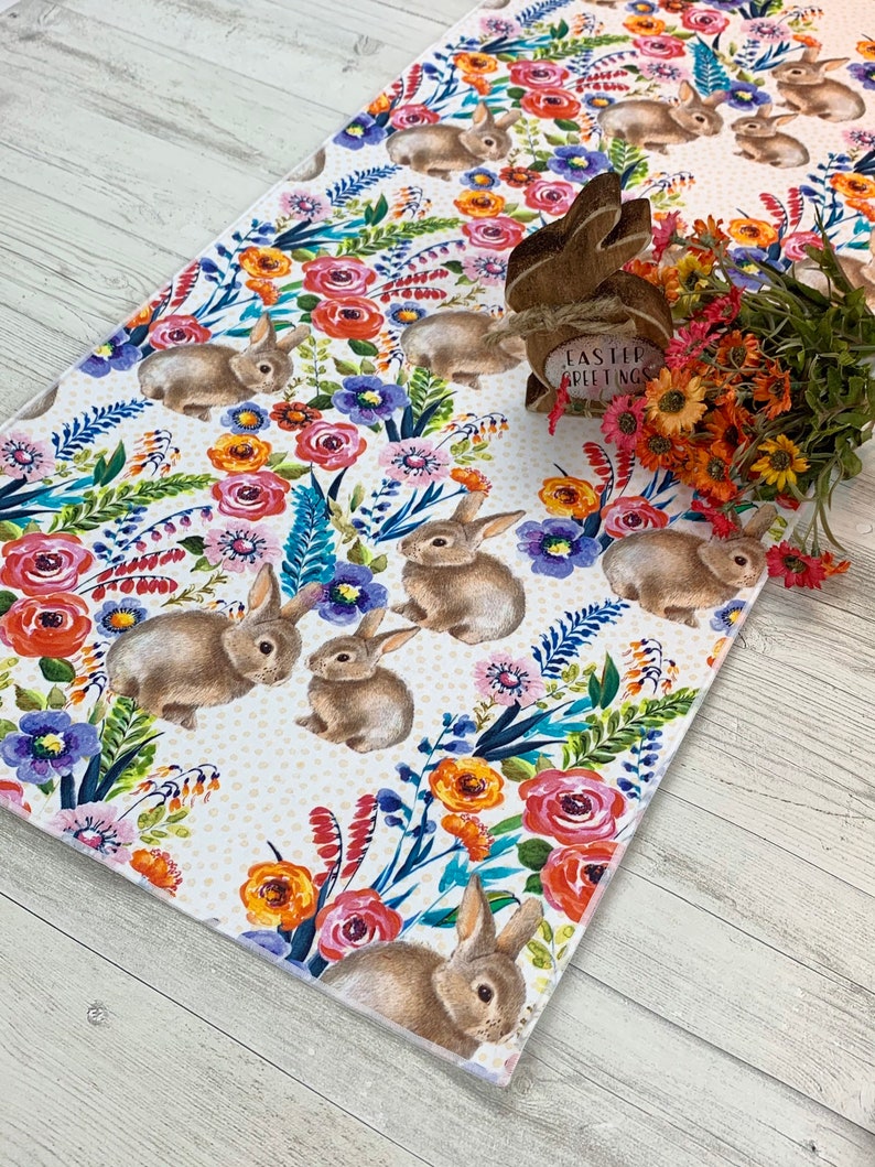 May include: A white table runner with a floral and bunny pattern. The runner features pink, red, orange, yellow, blue, and green flowers. The bunnies are brown and white. The text "Easter Greetings" is on a wooden bunny figurine.