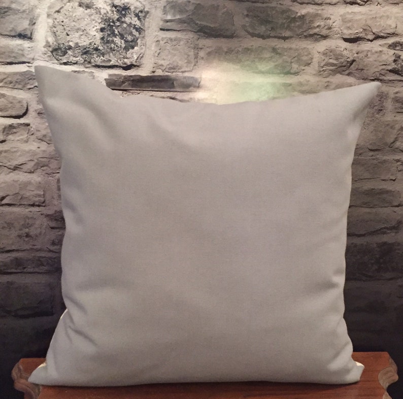 Puede incluir: Una almohada de tiro de color gris claro liso con un borde blanco. La almohada est&aacute; sobre una superficie de madera.