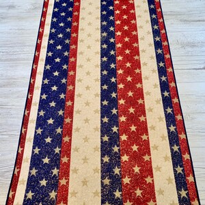May include: A red, white, and blue striped runner with a glittery finish. The runner features a repeating pattern of stars.