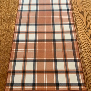 May include: A plaid table runner in shades of brown, white, and black. The runner has a subtle sheen and a woven texture.