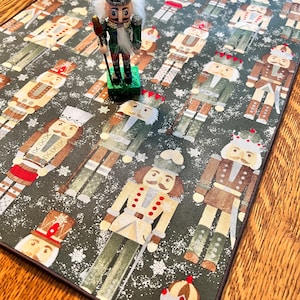 May include: A green table runner with a repeating pattern of nutcracker soldiers. The nutcracker soldiers are wearing different colored uniforms and hats. The runner has a brown border and a white snowflake pattern.