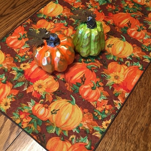 May include: A brown table runner with a pumpkin pattern. The runner is decorated with green and orange ceramic pumpkins.