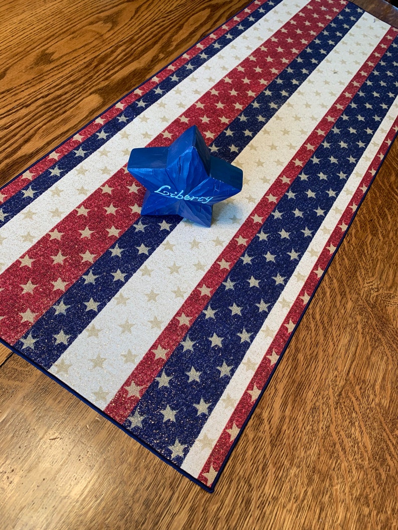 May include: A red, white, and blue table runner with a glittery finish. The runner features a repeating pattern of stars and stripes. A blue star-shaped ornament with the word "Liberty" written on it is placed on the runner.
