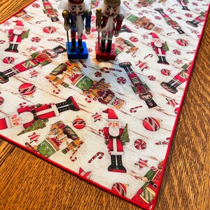 May include: A white table runner with a red border features a repeating pattern of nutcrackers, candy canes, and ornaments. Two nutcracker figurines are placed on the runner.