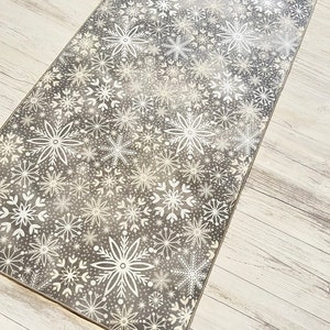 May include: A gray and white runner rug with a snowflake pattern. The rug has a distressed look and is perfect for adding a touch of winter charm to your home.