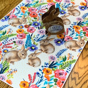 May include: A white table runner with a floral pattern featuring pink, orange, yellow, and blue flowers. The runner is decorated with brown bunnies and has a small wooden bunny cutout with the text "Easter Greetings" on it.