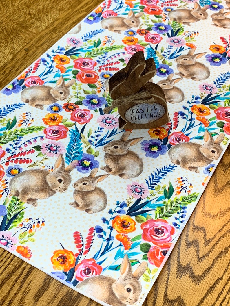 May include: A white table runner with a floral pattern featuring pink, orange, yellow, and blue flowers. The runner is decorated with brown bunnies and has a small wooden bunny cutout with the text "Easter Greetings" on it.