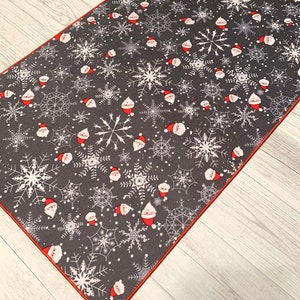 May include: A gray and white Christmas themed runner rug with a red border. The rug features a repeating pattern of snowflakes and Santa Claus faces.