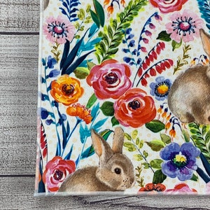May include: A white fabric with a floral pattern featuring pink, orange, and red flowers, green leaves, and two brown rabbits. The background is a pale yellow with white polka dots.