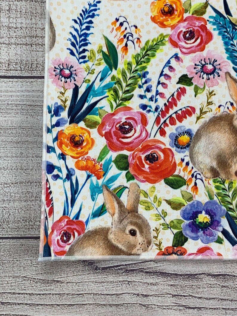 May include: A white fabric with a floral pattern featuring pink, orange, and red flowers, green leaves, and two brown rabbits. The background is a pale yellow with white polka dots.