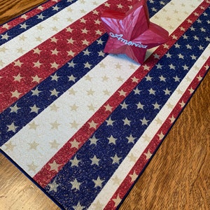 May include: A red, white, and blue table runner with a glittery finish. The runner features a repeating pattern of stars and stripes. A red wooden star with the word "America" is placed on top of the runner.