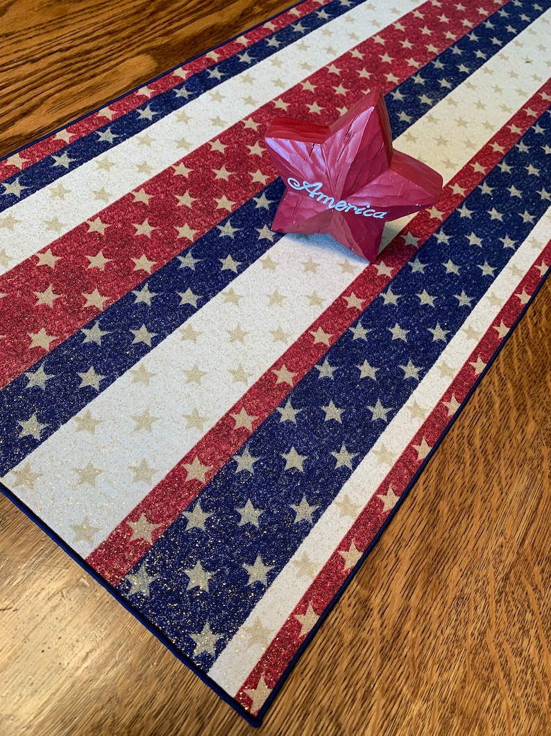 May include: A red, white, and blue table runner with a glittery finish. The runner features a repeating pattern of stars and stripes. A red wooden star with the word "America" is placed on top of the runner.