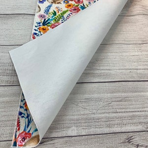 May include: A white table runner with a floral and bunny design. The runner is folded in half to show the white backing.