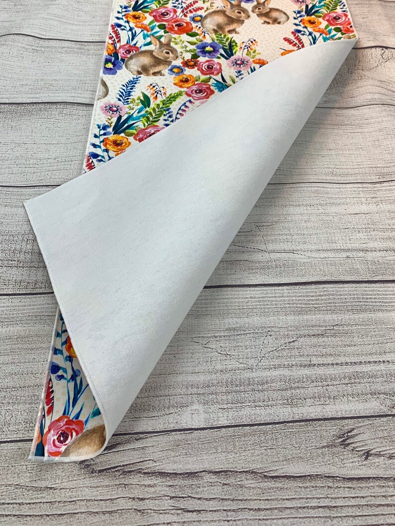 May include: A white table runner with a floral and bunny design. The runner is folded in half to show the white backing.