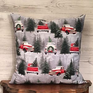 May include: A gray throw pillow with a red truck and Christmas tree pattern. The truck is carrying a Christmas tree and is driving through a snowy forest.